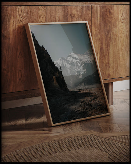 Ein gerahmtes Poster von einem breiten Flussbett in den Dolomiten, umgeben von schneebedeckten Bergen, an einer Holzkommode angelehnt und stehend auf dem Holzboden