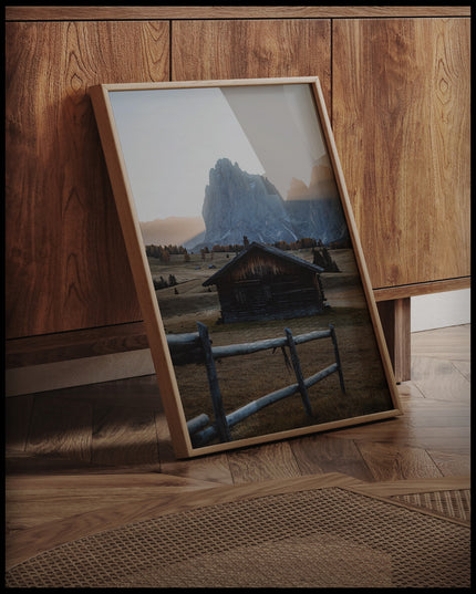 Ein gerahmtes Poster von einer rustikalen Hütte in einer herbstlichen Berglandschaft, an einer Holzkommode angelehnt und stehend auf dem Holzboden