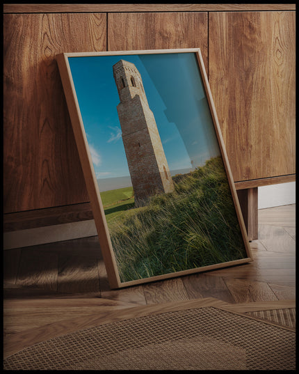 Ein gerahmtes Poster von einem steinernen Turm auf einer grünen, hügeligen Wiese, an einer Holzkommode angelehnt und stehend auf dem Holzboden