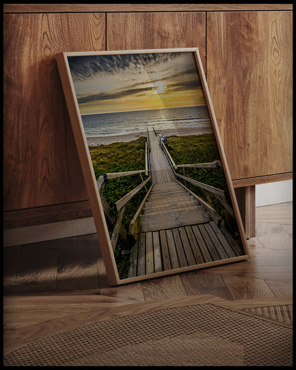 Ein gerahmtes Poster von einem Holzsteg mit einer Treppe, der durch Dünen zum Strand führt, an einer Holzkommode angelehnt und stehend auf dem Holzboden