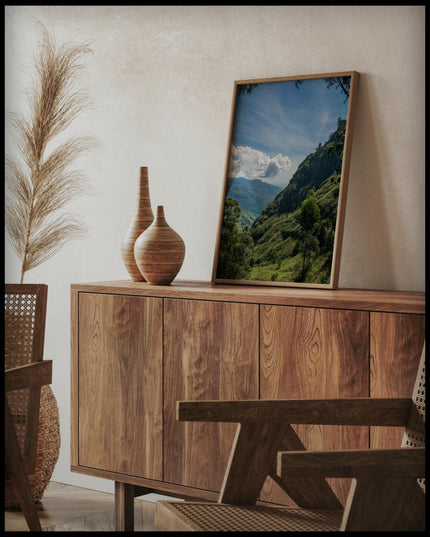 Ein gerahmtes Poster einer bewaldeten Berglandschaft in einer tropischen Region, auf einer Holzkommode an die Wand angelehnt