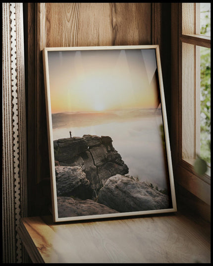 Ein gerahmtes Poster von einem Bergsteiger, der bei Sonnenaufgang auf einer Klippe steht, vor einem Fenster an die Wand angelehnt