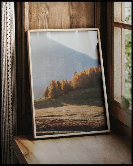 Ein gerahmtes Poster von einer Herbstlandschaft in den Dolomiten mit Bäumen im Sonnenlicht, vor einem Fenster an die Wand angelehnt