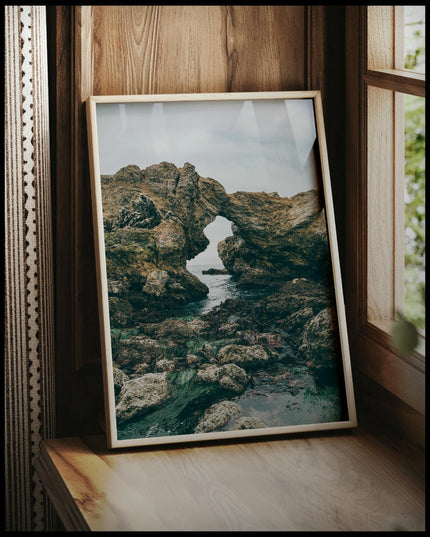 Ein gerahmtes Poster von einer felsigen Küste mit einer natürlichen Felsformation, die ein Fenster zum Meer bildet, vor einem Fenster an die Wand angelehnt