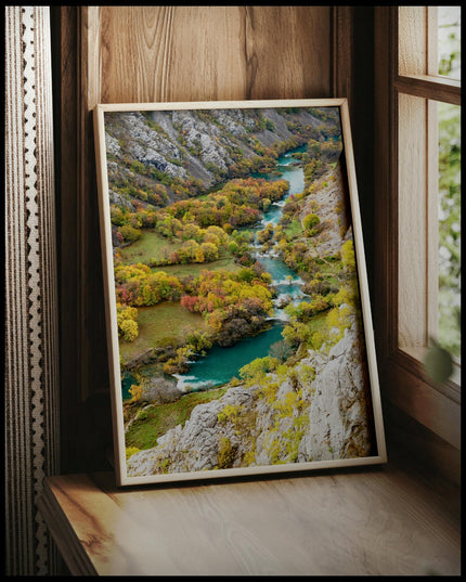 Ein gerahmtes Poster von einem Fluss der sich, umgeben von mehreren Büschen, durch eine Berglandschaft erstreckt, vor einem Fenster an die Wand angelehnt