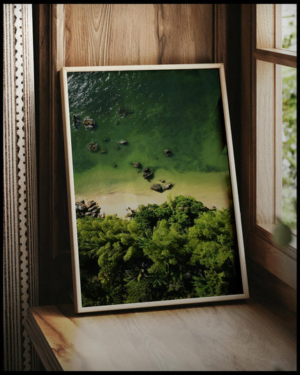 Ein gerahmtes Poster von einem Sandstrand mit grünlichem Wasser vor einem tropischen Wald, vor einem Fenster an die Wand angelehnt