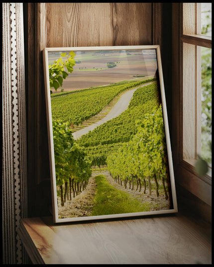 Ein gerahmtes Poster von einer Straße, die durch hellgrüne Weinberge führt, vor einem Fenster an die Wand angelehnt